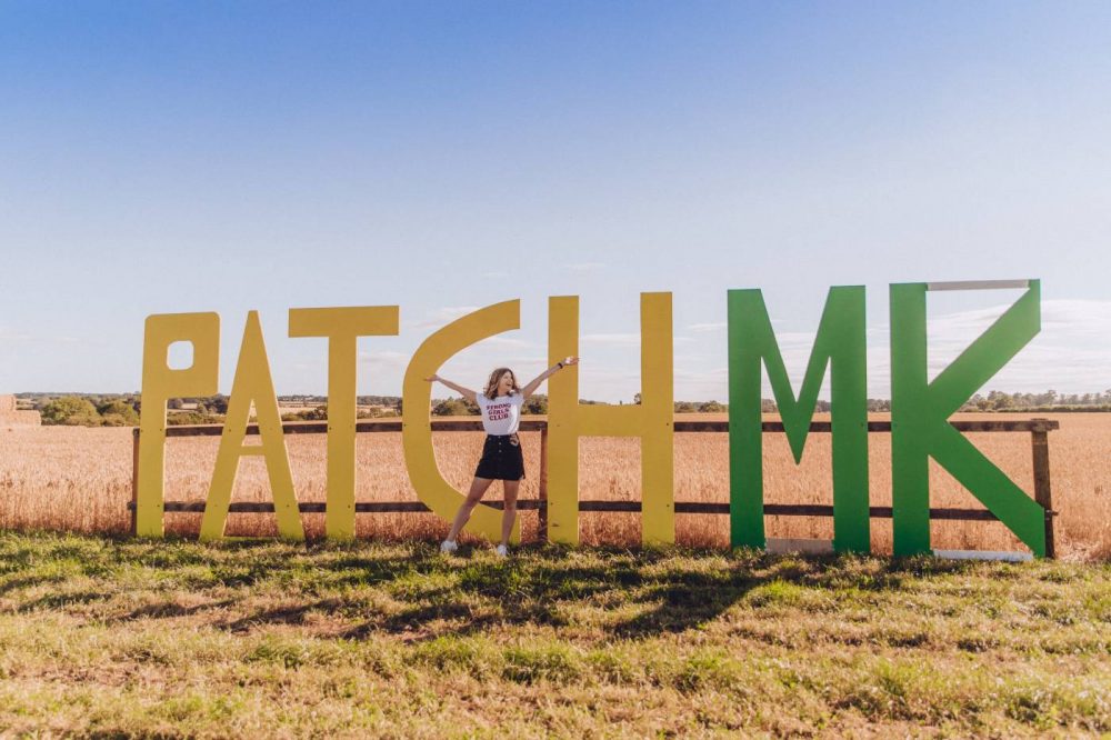 Sophie etc standing in a field in front of a sign that says 'Patch MK'