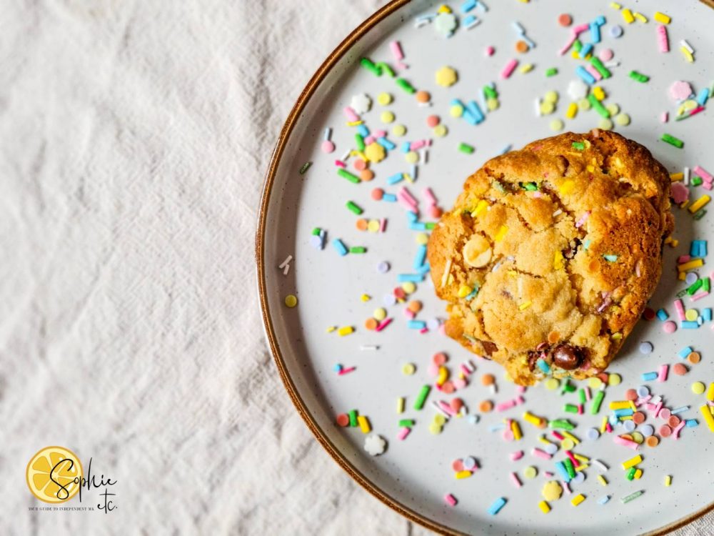 A chocolate chip and rainbow sprinkel NYC style cookie on a plate of sprinkles. 